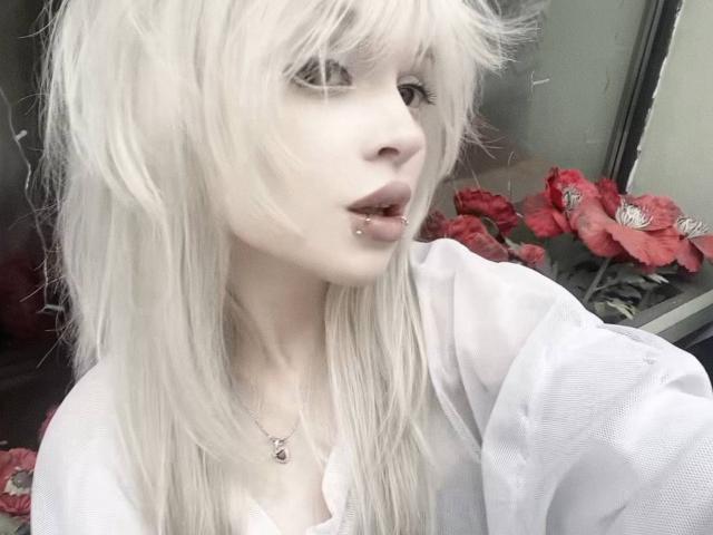 HortenciaVergin, con la pelle chiara, i capelli biondo platino e un piercing al labbro, indossa una maglietta bianca, guarda di lato, mostrando il suo seno piccolo e una delicata collana..