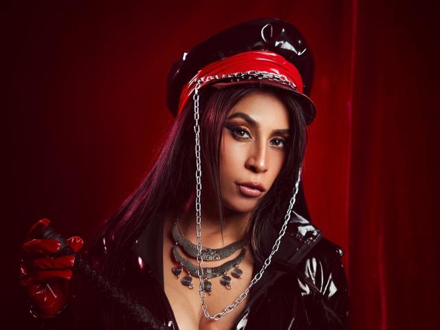 AfroditaDoir with long black hair, red eyes, and glossy black latex, wearing a red and black cap with chains, holding a black cat-o'-nine-tails. Red lips, necklace.