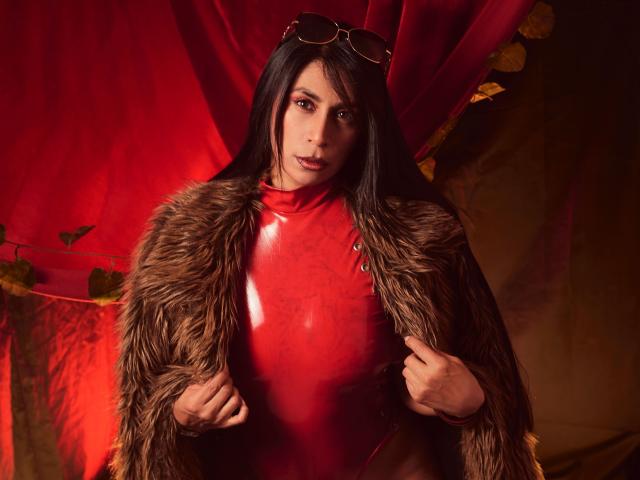 AfroditaDoir mit langen, schwarzen Haaren, die ein glänzendes rotes Latexoberteil und einen braunen Pelzmantel trägt, wobei der Mantel offen gehalten ist, und mit Aviator-Sonnenbrillen auf dem Kopf..