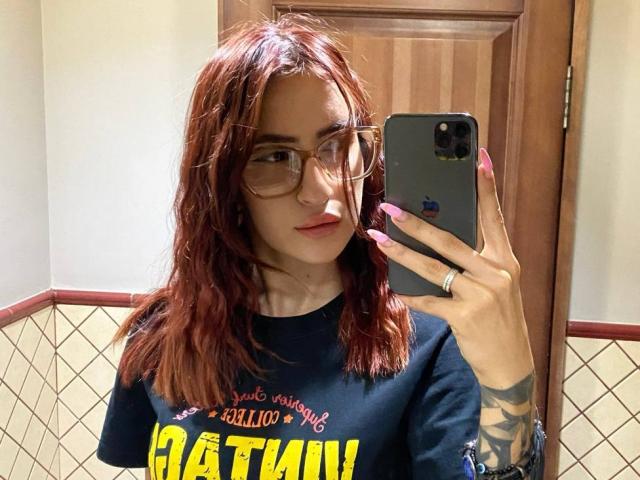 Precious Lambrecht, com cabelo ruivo, óculos e unhas cor-de-rosa, segurando um telefone, vestindo uma camiseta preta da CATAN, mostrando uma tatuagem no braço direito, em um banheiro..