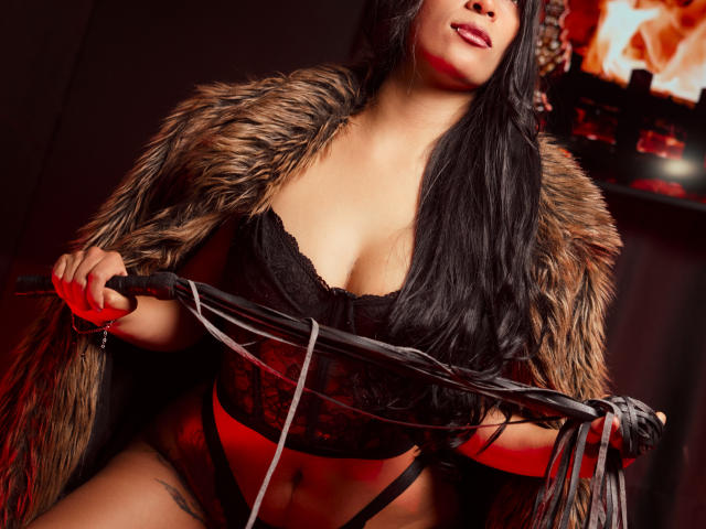 AfroditaDoir with long black hair, wearing a fur coat, black lace lingerie, and holding a whip, showing her tits and red garter, in a sexy, dark room.