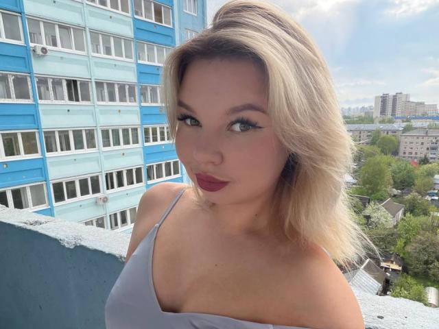 ChuHeal tiene cabello rubio, ojos azules y lápiz labial rojo, y lleva una blusa blanca con tirantes finos, mostrando su busto y un rostro lindo, de pie en un balcón con un edificio azul..
