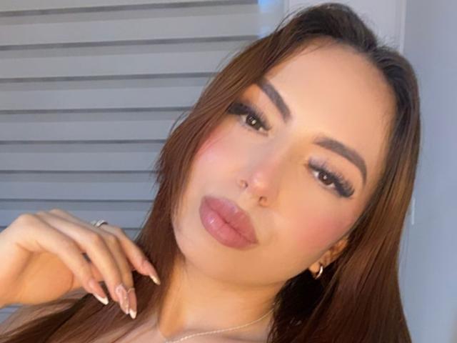 Ivonne Falcone hat braunes Haar, volle, rosafarbene Lippen und dunkle Augenbrauen. Sie schaut mit leichtem Schmollmund in die Kamera, ihre rechte Hand berührt ihr Haar..