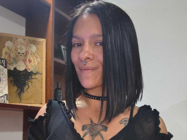 AmelieBlake has black shoulder-length hair, a nose ring, a choker, and a black top showing a tattoo of a spider on her chest. She's smiling.