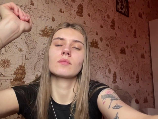 DeliciaQuanstrum, con los ojos cerrados, camisa negra y cabello rubio largo, levantando el brazo izquierdo, mostrando un tatuaje en el brazo derecho, en una habitación con una pared con un diseño de mapa..