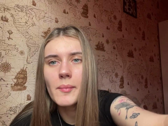 DeliciaQuanstrum tem cabelos loiros longos, olhos verdes e uma tatuagem no braço direito que mostra um pássaro colorido. Ela está vestindo uma camisa preta e tem um sorriso discreto..