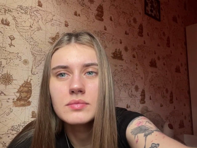DeliciaQuanstrum, con lunghi capelli biondi, pelle chiara e un tatuaggio sul braccio destro, che appare seria con le labbra rosate, indossa una maglietta nera..