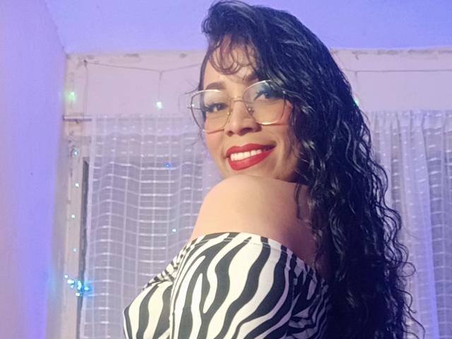 FetishLoverAna with curly black hair, glasses, red lipstick, and zebra-print dress, smiling at camera, showing bare shoulder, wearing glasses, looking sexy.