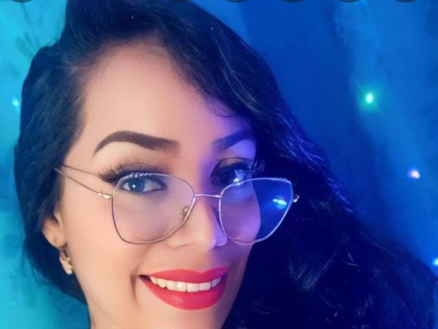 FetishLoverAna, avec du rouge à lèvres, des cheveux noirs ondulés et des lunettes, souriant à la caméra, avec un teint bleuté, portant des boucles d'oreilles, et ayant l'air mignonne et sexy..