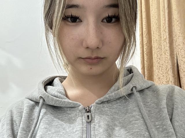 Amiee Shibata heeft een schattig, sproetig gezicht met lange wimpers, draagt een grijze hoodie, haar blonde haar zit in een rommelige knot, en ze heeft een klein sproetje op haar kin..