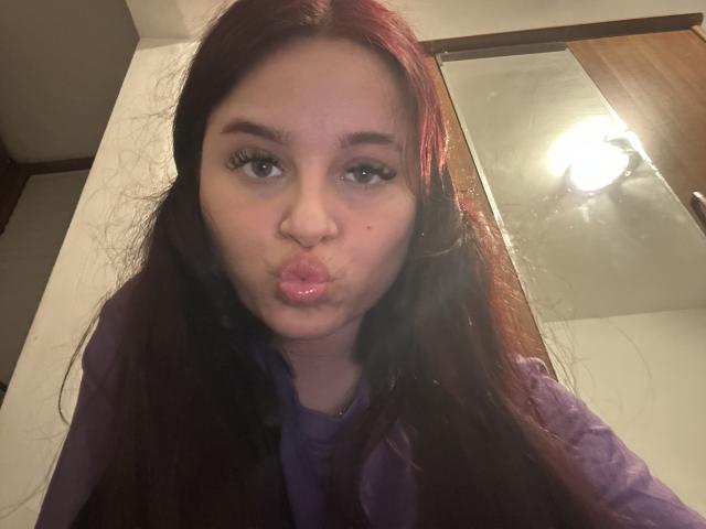 Lucie, met lang rood haar, volle roze lippen en een klein neusje, draagt een paars shirt en heeft een schattelijk, aantrekkelijk gezicht met subtiele make-up..