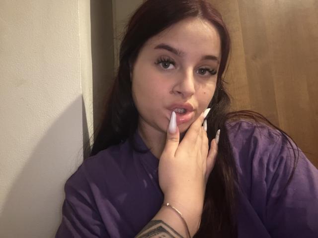 Lucie, met lang bruin haar, een paars shirt en lange witte nagels, die aan haar vinger knabbelt en een tatoeage op haar rechterpols laat zien. Haar lippen zijn lichtjes open..