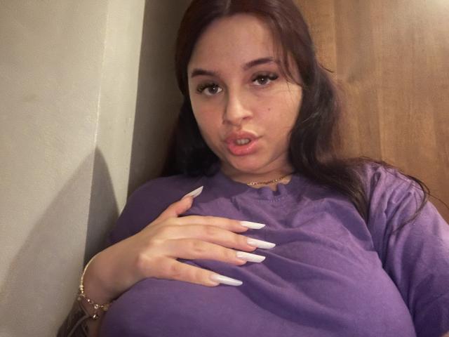 Lucie, met lang bruin haar, volle lippen en een paars shirt, die haar grote borsten aanraakt met witte, lange nagels, en die in de camera kijkt. Ze draagt een gouden armband en ketting..