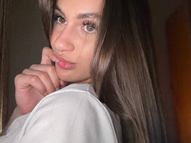 EllenKienbaum with brown eyes, pink lips, and long brown hair, wearing a white ribbed shirt, touching her chin with a finger, looking at the camera.