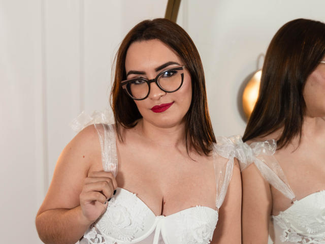 Lindsey Ford con gli occhiali, rossetto rosso e vestito bianco in pizzo, che mostra il seno, tiene una cinghia, in piedi di fronte a uno specchio, capelli castani, riflesso, sorriso dolce, formosa, carina..