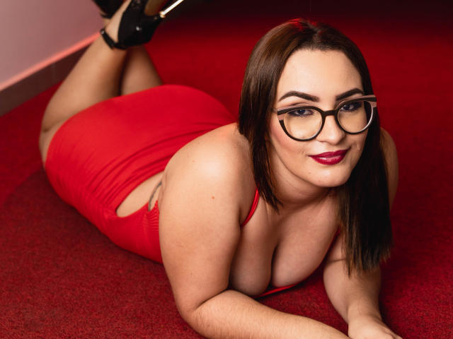 Lindsey Ford, con il seno prosperoso e un vestito rosso, è sdraiata su un tappeto rosso, con le gambe sollevate, indossando tacchi neri, occhiali e rossetto rosso, e sorridendo alla macchina fotografica..