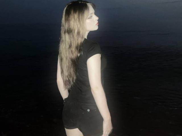HortenciaVergin with long brown hair, black t-shirt, and black shorts, standing with her back to the camera, looking to the side, in a dark, illuminated night setting.