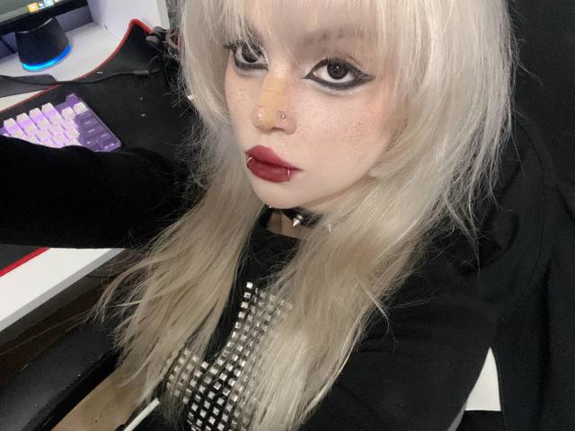 HortenciaVergin with platinum blonde hair, black eyeliner, red lipstick, nose piercing, bandage on nose, black shirt with silver studs, sitting at a computer, looking at camera.
