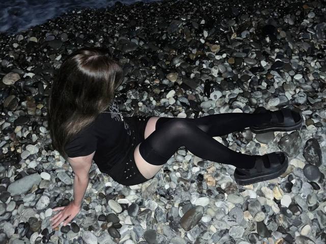 HortenciaVergin, avec de longs cheveux bruns, un t-shirt noir, un short noir, des bas noirs montants jusqu'aux cuisses et des baskets noires, assise sur une plage rocheuse, assise de manière à ce que ses fesses soient en l'air, les jambes écartées, les bras derrière elle..