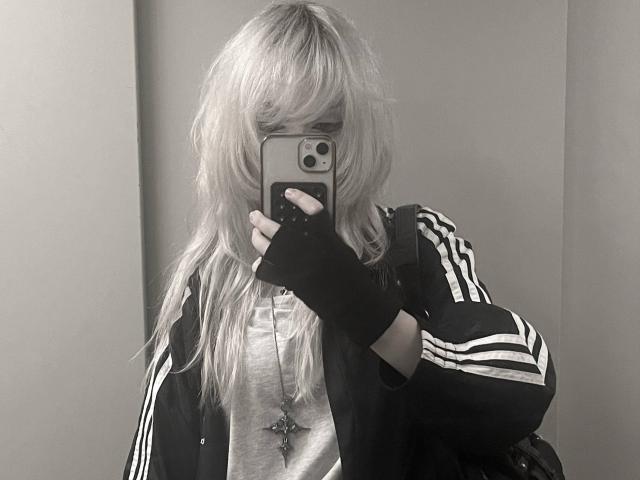 HortenciaVergin, avec ses cheveux blonds et ondulés, portant une veste Adidas noire, un t-shirt gris, des gants noirs sans doigts, prenant un selfie devant un miroir, son téléphone cachant son visage, avec un mur blanc en arrière-plan..