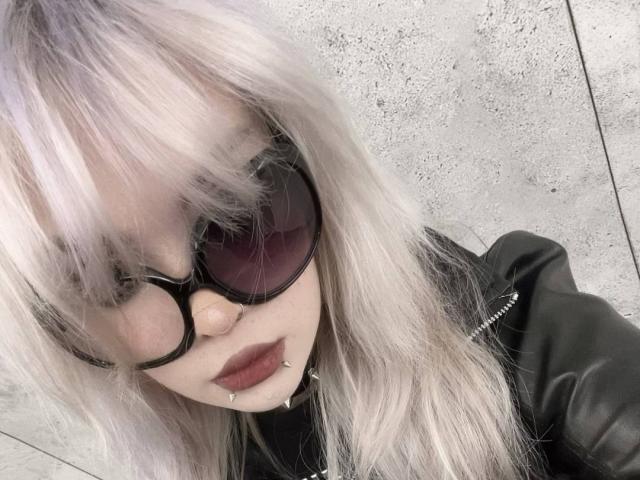 Hortencia Vergin mit hellrosa-weißem Haar, schwarzen Sonnenbrillen, dunklem Lippenstift, einer schwarzen Lederjacke und Nasenpiercings, die nach unten schaut. Ihre Brüste sind nicht sichtbar. Eine Betonmauer im Hintergrund..