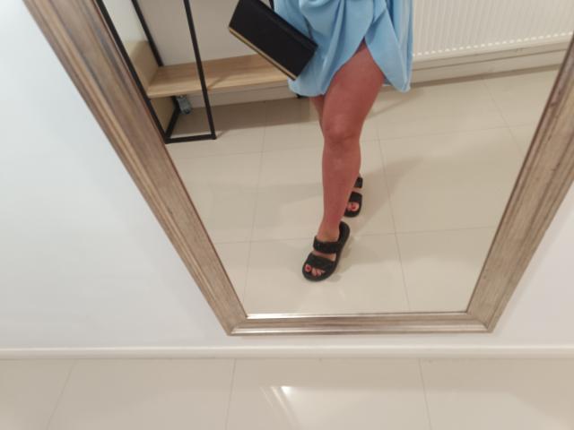 AmberSunshine's reflection shows her in a blue dress, black sandals with straps, and a black clutch. Her bare, tan, and slightly muscular legs are visible.