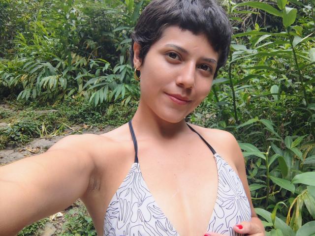 Scarlett69Hot with short black hair, small black earrings, wearing a white and gray patterned bikini top, showing her small tits and armpit hair, standing in a green jungle.