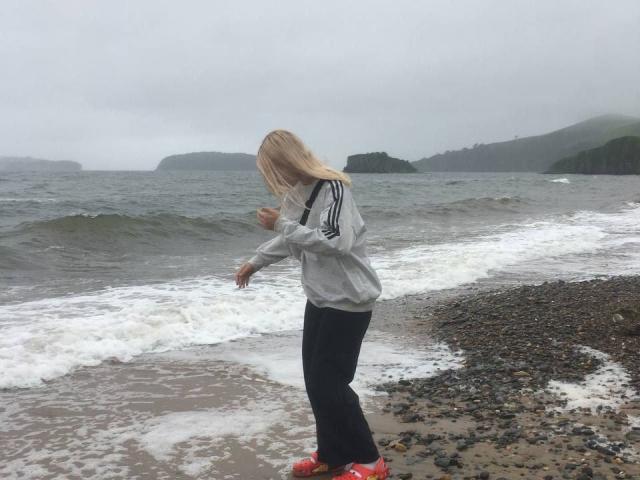 AnnamaeSvehla with blonde hair, grey hoodie, black pants, and red shoes, standing on a pebbly beach, facing the ocean, with small waves and cloudy sky.