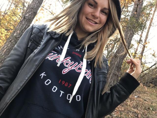 BelleLisaG con cabello rubio, sonriendo, vistiendo una chaqueta de cuero negra y una sudadera negra con la palabra "Trophy", sosteniendo su cabello, de pie en un bosque..