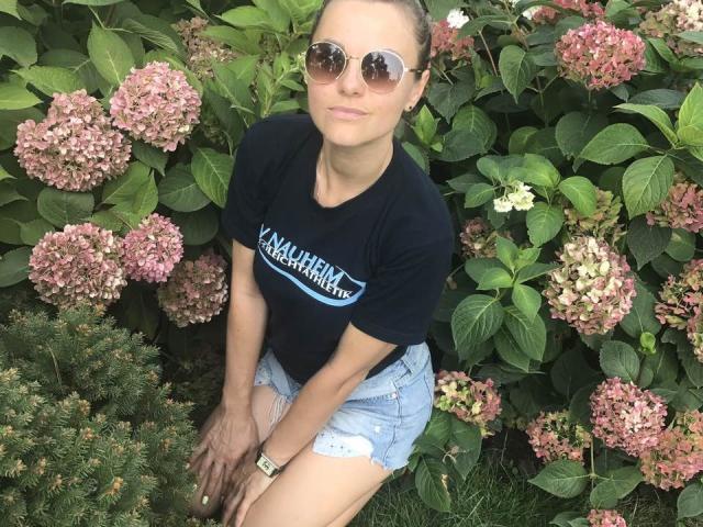 BelleLisaG, con piel morena, cabello castaño recogido en un moño, usando gafas de sol redondas, una camiseta negra y pantalones cortos vaqueros, sentada frente a unas hortensias rosas y verdes..