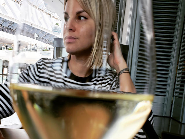 BelleLisaG avec une coupe au carré blonde, une chemise rayée, une main dans les cheveux, regardant sur le côté, verre flou au premier plan, intérieur, décontracté, expression concentrée, bracelet au poignet droit..