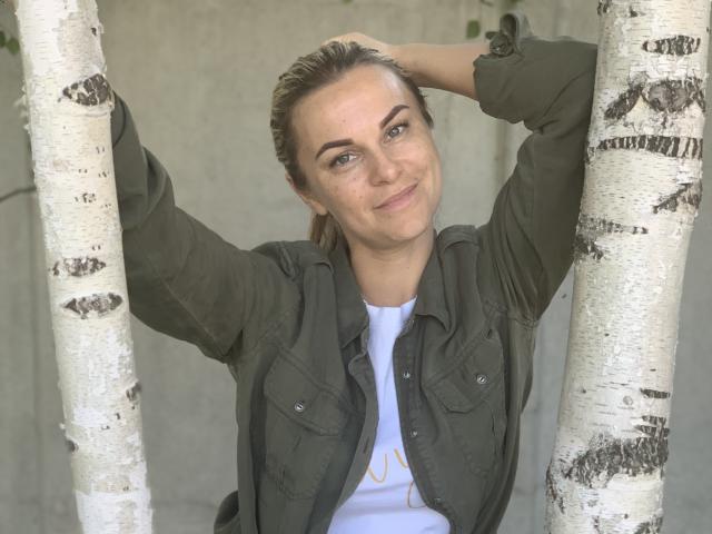 BelleLisaG con los brazos levantados, sosteniendo árboles de abedul blancos, vistiendo una chaqueta verde oliva sobre una camisa blanca, sonriendo sutilmente, cabello castaño en una coleta, con pecas claras en la cara..