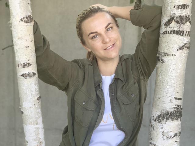 BelleLisaG with arms raised, smiling, wearing an olive green shirt and white tee, standing between two birch trees, brown hair pulled back, green eyes, light skin, and a wristwatch.