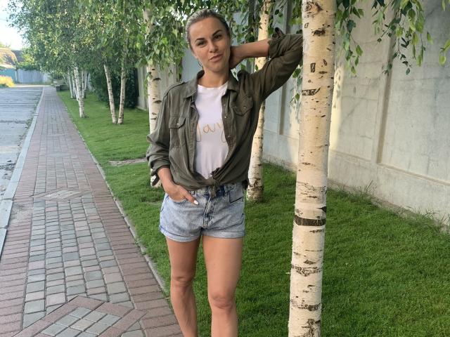 BelleLisaG with tied green shirt, white tee, and blue denim shorts, standing by birch tree, hand on head, green grass, brick sidewalk, and gray wall in background.