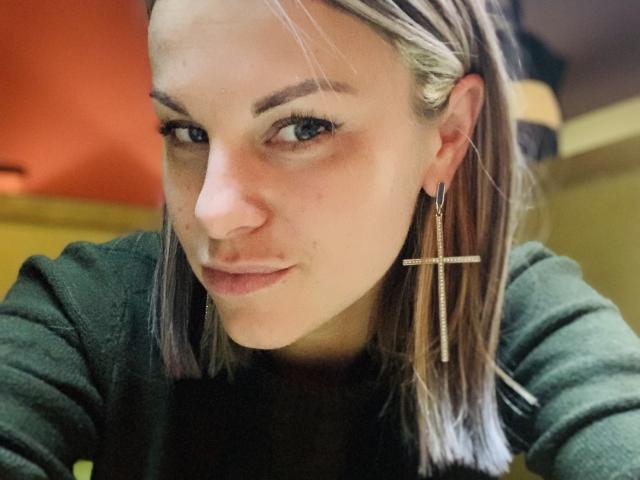 BelleLisaG, avec ses cheveux blonds lisses, ses yeux verts, portant un haut vert et de grandes boucles d'oreilles en forme de croix, a une légère moue et un petit bouton visible sur sa joue..
