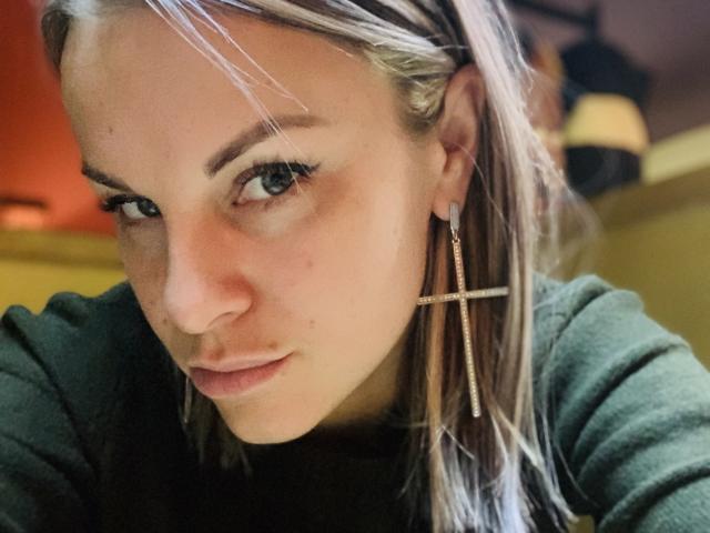 BelleLisaG, con i capelli biondi, gli occhi verdi e un'espressione seria, indossa una camicia verde e un orecchino a forma di croce. Il suo viso è leggermente arrossato..