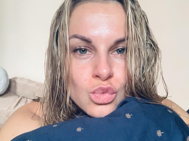 BelleLisaG con capelli biondo platino umidi, labbra rosa gonfie e una camicia blu con piccoli motivi bianchi, che guarda direttamente nella fotocamera con le sopracciglia leggermente alzate. Pelle chiara, lentiggini leggere e occhi azzurri..