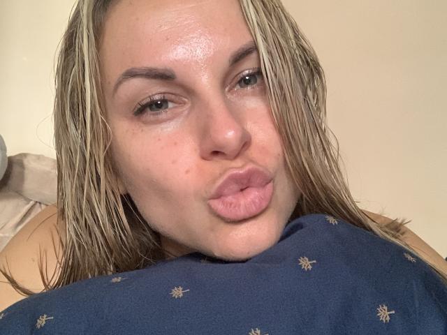 BelleLisaG con i capelli biondo platino umidi, labbra rosa gonfie e lentiggini, che fa una smorfia, indossa una camicia blu con piccoli motivi dorati. I suoi occhi sono socchiusi..