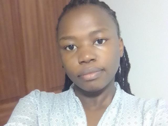 Yollah has dark skin, small nose, and full lips. She's wearing a white patterned shirt with black braided hair. Her eyes are looking at the camera.