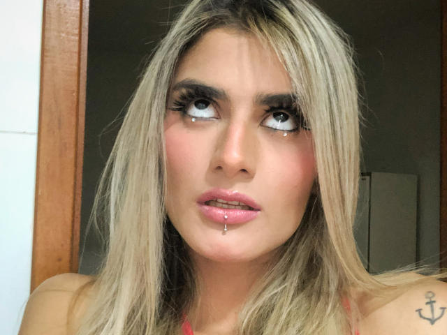 A BabeZina tem cabelo loiro, lábios rosados e brilhantes com uma gota de saliva, e uma pequena tatuagem de âncora no ombro direito. Ela está olhando para o lado com olhos grandes..