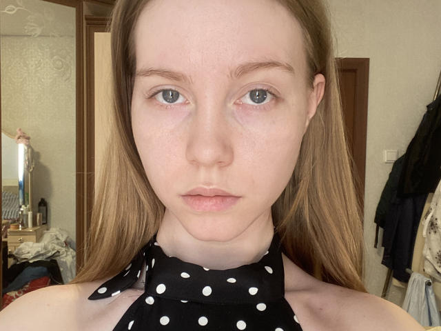 LianeResper, com pele clara, olhos azuis e cabelo castanho liso, vestindo uma blusa preta com bolinhas, com uma expressão séria e um leve franzir de lábios, em um quarto bem iluminado..