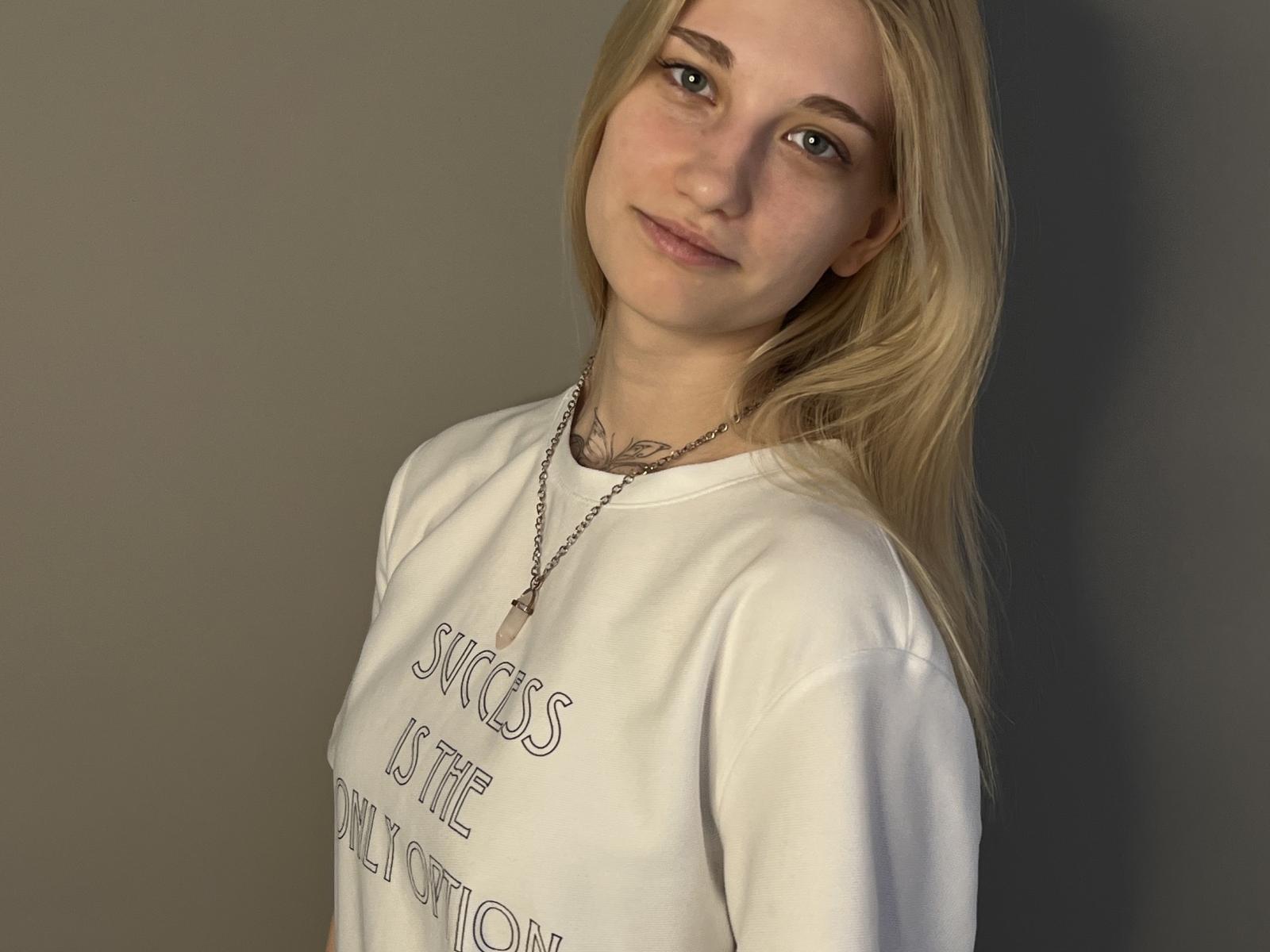 LuciTisue with blonde hair, pale skin, and subtle makeup, wearing a white shirt and necklace, has a small tattoo on her neck and a slight, confident smile.