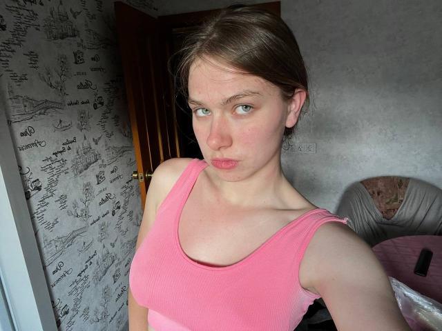 ShiraWolny, con labios carnosos, vestida con una camiseta rosa de tirantes y textura vertical, mostrando sus pequeños senos y sus delgados brazos, cabello castaño, ojos verdes, en una habitación con poca luz..