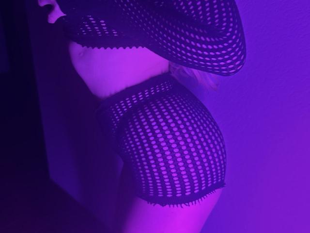 BalletSwan in una posa illuminata di viola, indossa lingerie nera a rete con il seno coperto, mostrando il sedere con pantaloncini a rete coordinati, in piedi di profilo, con le braccia alzate..