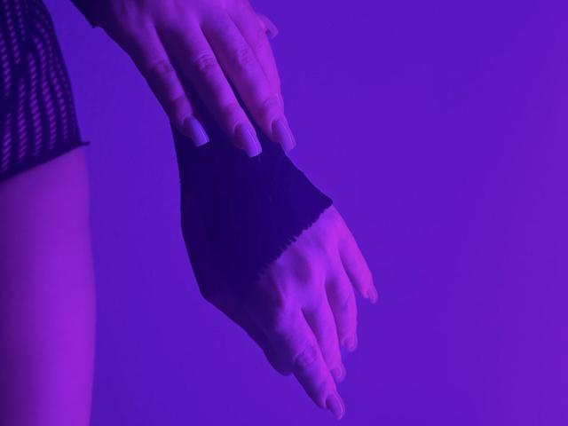 Las manos de BalletSwan, que lleva un guante negro sin dedos, están colocadas contra un fondo iluminado en color púrpura. Sus uñas son largas y blancas. Su muslo, cubierto con medias de red, es parcialmente visible..