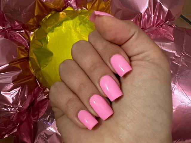 La main de BalletSwan, avec des ongles roses brillants, tenant une balle jaune et froissée, sur du papier rose froissé. Les seins ne sont pas visibles. L'attention est portée sur la main et les ongles..