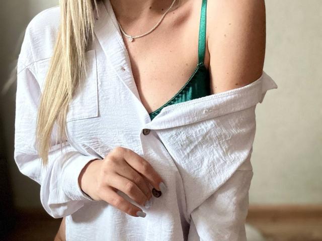 BalletSwan con una camisa blanca de botones, llevada de forma que deja un hombro al descubierto, mostrando un sujetador verde y el escote, cabello rubio largo y un collar de plata, sosteniendo la camisa con su mano..