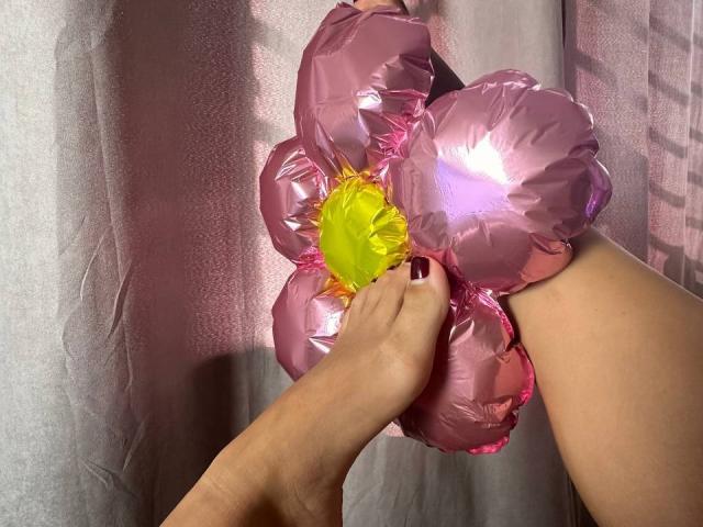 Las manos de BalletSwan sostienen delicadas flores en forma de globos rosas con un centro amarillo, sus uñas pintadas de rojo oscuro, y una parte de su pierna es visible, sobre un fondo suave, iluminado en tonos rosados y con textura..