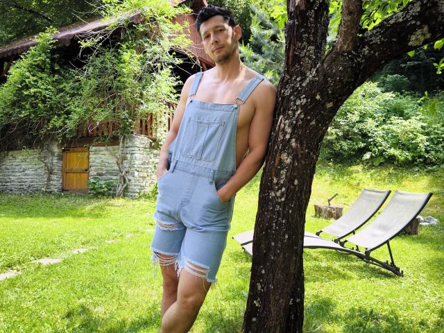 BrutusK, avec une carrure musclée, se tient appuyé contre un arbre, vêtu d'une salopette en jean bleu déchirée, les mains dans les poches. On aperçoit de l'herbe verte et une chaise longue en bois en arrière-plan..