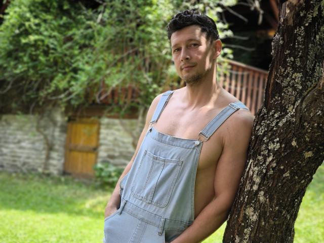 BrutusK leunt tegen een boom, zonder shirt, in grijze denim overalls, en laat een getraind, gespierd rug en armen zien, met een lichte, zelfverzekerde glimlach. Zijn achterkant is niet zichtbaar..